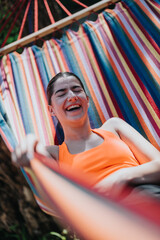 A cheerful woman rests in a vibrant, rainbow-colored hammock under sunlight, enjoying a pleasant outdoor ambiance.