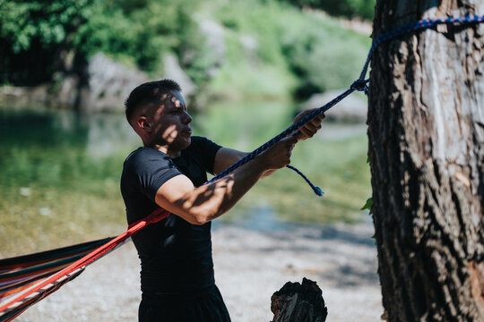 Outdoor enthusiast tightening a hammock line by a tree near a peaceful riverside setting.