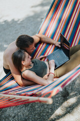 A couple works together on a laptop while reclining in a colorful hammock outdoors, enjoying...
