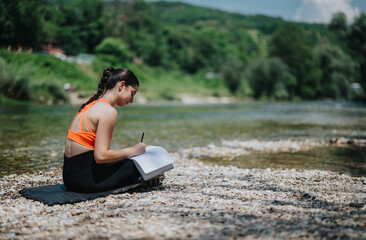 A young woman sits by the river on a sunny day, writing in her journal. The tranquil surroundings...