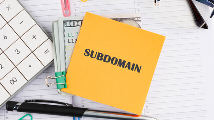 A desk setup features a bright yellow note labeled subdomain along with a stack of cash, a calculator, and various office supplies, indicating financial planning or organization