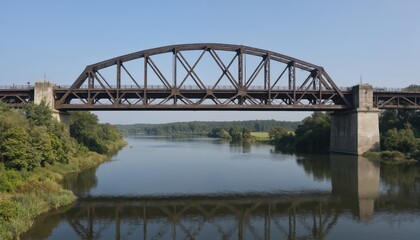 Naklejka premium Majestic steel truss bridge over calm river. Towering structure with robust brown paint, cables anchored in ground. Water below mirrors bridge design. Trees dot landscape, vibrant green leaves. Clear
