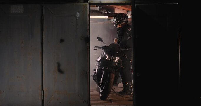Kyiv, Ukraine 24.08.2023  View of the garage at night through the opened door. A guy in motorcycle gear puts on a helmet, gets on a motorcycle and  ready to ride, turning on the headlight