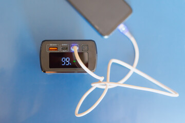 Power bank with 99 percent numbers on display and white cable in port charging phone close-up blue background, autonomous power
