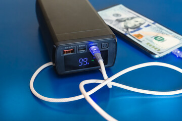 Power bank with 99 percent numbers on display and white cable in port charging phone with hundred dollars on it close-up blue background, autonomous power