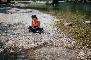 A woman, dressed in active wear, sits on a pebbled riverbank writing in her journal, surrounded by calm waters and natural scenery.