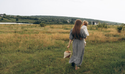 A young woman in a checkered dress carries a child while walking in a serene countryside landscape.
