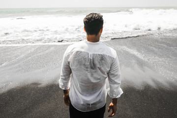 Man in White Shirt Standing on Beach Facing Ocean Waves.