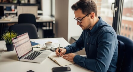 Young man wearing glasses and a blue shirt writing in a notebook at his desk with a laptop and coffee