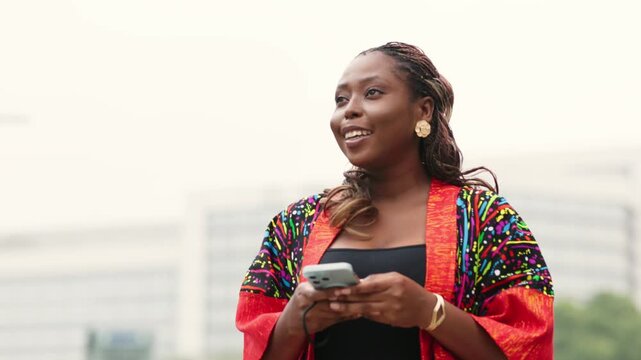 Smiling Black woman in vibrant African print kimono using smartphone outdoors.