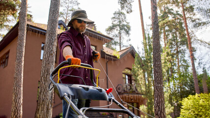 Man Using a Lawn Mower Outside a Large Home Surrounded by Pine Trees