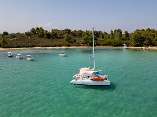 Catamaran anchored in crystal clear turquoise water near a forested Greek island beach, surrounded...