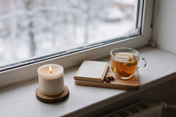 Winter wellness flat lay with immunity tea, ginger, clove, aroma candle on wooden coaster, textured journal and snowy blurred window in soft natural calming tones