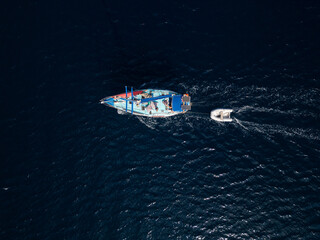 Aerial top view of a colorful sailboat towing a small dinghy through deep blue sea water. The...