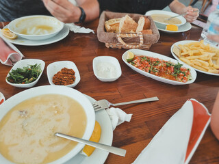 Table set with Turkish bread, various sauces, fries, and soups, with visible hands of a man and a woman reaching for food. Casual dining scene.
