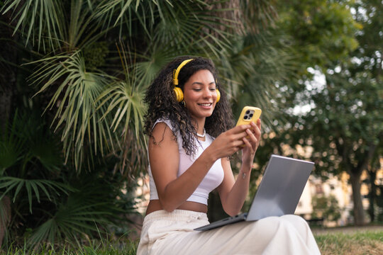 Young woman working remotely in a park at sunset, using a laptop and smartphone