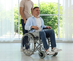 Caregiver with man in wheelchair near window indoors, closeup