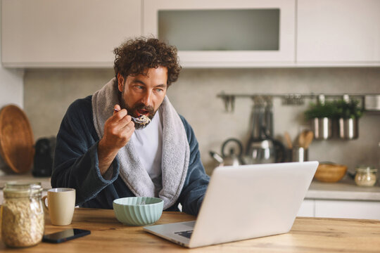 A man sits at a wooden table, wearing a bathrobe, eating cereal from a bowl while focused on a laptop screen in a bright kitchen decorated with simple elements.
