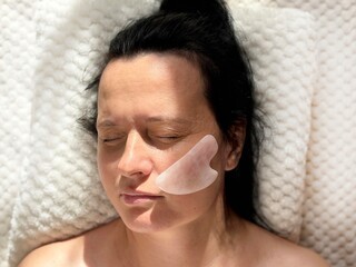 Mature woman lying on cosmetology table during professional facial massage with natural scraper....