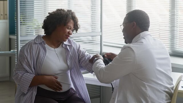 Medium shot of African American healthcare professional checking pregnant patient blood pressure using cuff and monitor, ensuring vital signs during prenatal medical appointment