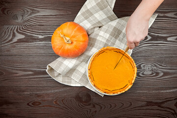 Hand slicing into a freshly baked pumpkin pie on a wooden table, with a vibrant orange pumpkin and...
