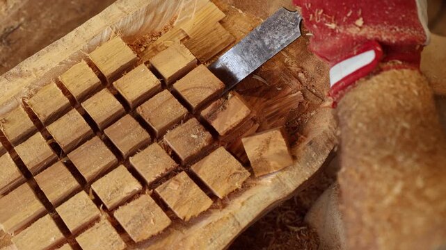 A craftsman wearing gloves carefully carves a wooden block using a chisel and mallet. The scene captures the precision and rhythm of traditional woodcraft artistry