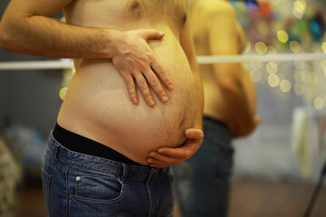 Pregnant Man Holding Belly in Jeans in Front of Mirror with Lights in Background