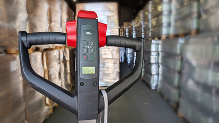 Electric pallet jack with control panel in a warehouse, surrounded by stacked pallets and goods,...