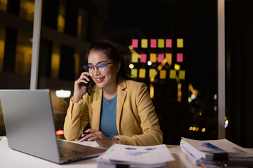 Asian female working on digital laptop with smartphone from work at night.