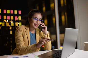 Asian female working on digital laptop with smartphone from work at night.