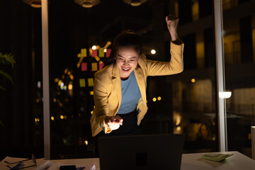 Excited young Asian woman with laptop at work desk in office a night time, feeling joy celebrating ...