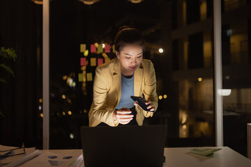 Asian female working on digital laptop with smartphone from work at night.