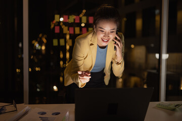 Asian female working on digital laptop with smartphone from work at night.