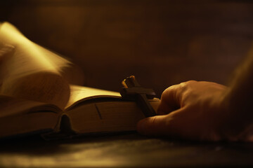 Hand Holding Cross Over Open Bible in Candle Light Creating Shadows for Devotional Reflection