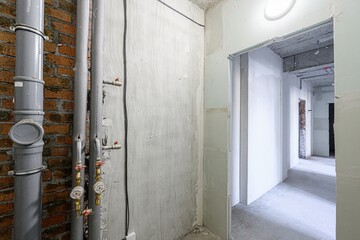 unfinished room with exposed brick, pipes, and concrete walls. A doorway leads to a hallway, revealing more unfinished spaces and infrastructure