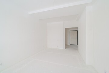 Bright, empty room with white walls and floor. A doorway leads to a hallway with a closed door at the end