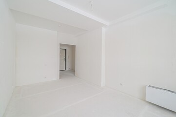 stark, white room in an unfinished state with a doorway leading to another room. A radiator is visible on the wall.