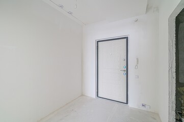 bright, empty room with white walls and a front door. The room appears to be undergoing renovation or is newly constructed
