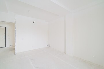 empty, all-white room under construction. Visible are walls, ceiling, a door, and some exposed piping and electrical outlets. The floor looks unfinished