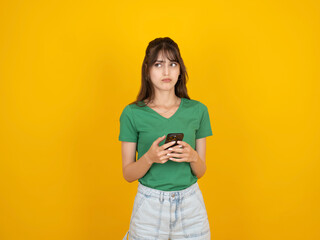 Confused young woman, photo of young caucasian 20s thinking confused young woman hold use mobile phone looking aside  purses lips being dissatisfied wears green t-shirt and jeans isolated on yellow.