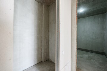 An unfinished room with bare concrete walls and floors, with a glimpse into another room through a...