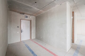 An unfinished room with bare concrete walls and floor. A door, covered in plastic, is in the left. Visible wiring and exposed brick are present