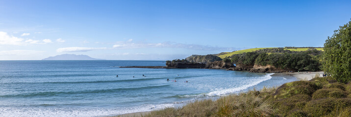 Surfers enjoy the waves at Tawharanui in New Zealand. The clear blue water and sky create a peaceful atmosphere for water sports and recreation. Coastal landscape.