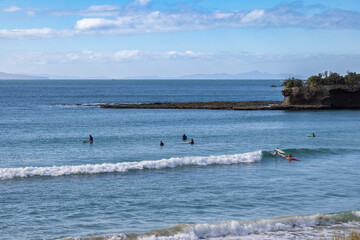Surfers enjoy the waves at Tawharanui Beach in Auckland, New Zealand. People are surfing and waiting for waves on a sunny day. The ocean is calm and blue.