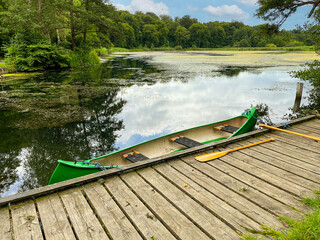 Scenic and idyllic depiction of an empty green multi-person canoe resting on a wooden platform by a lake with blue sky and white clouds. In the background a forest.