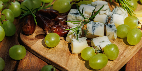 Wooden board with cheese cubes, grapes, rosemary and dates arranged as rustic summer appetizer..