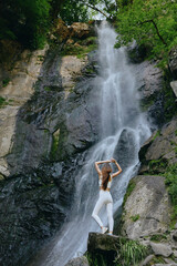 woman standing near waterfall in nature wearing white activewear, serene outdoor scene with rocky...