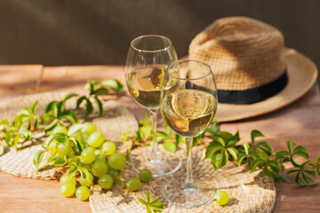 Two glasses of white wine with green grapes and straw hat on wooden table in natural sunlight..