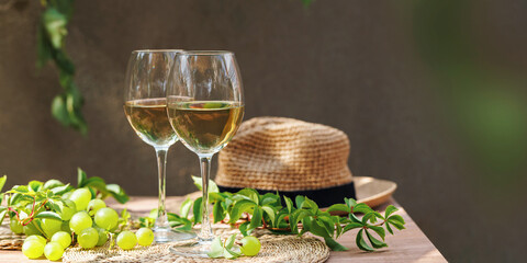 Two wine glasses and fresh grapes on summer patio with straw hat and green leaves..