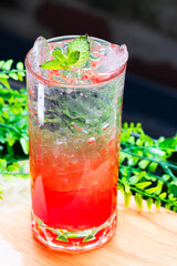 Refreshing red cocktail with ice and mint leaves served in a clear glass on a wooden surface with green foliage background.